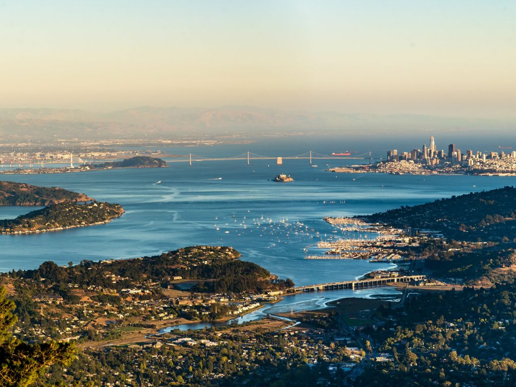 an aerial photo of the bay and surrounding coast line, with a suspension bridge in the middle, and a background of mountains and sky