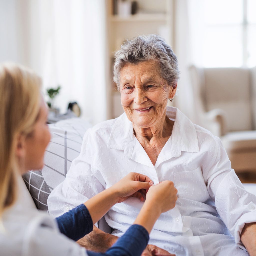 a care employee helps an elderly resident with their shirt; the setting is the resident's home