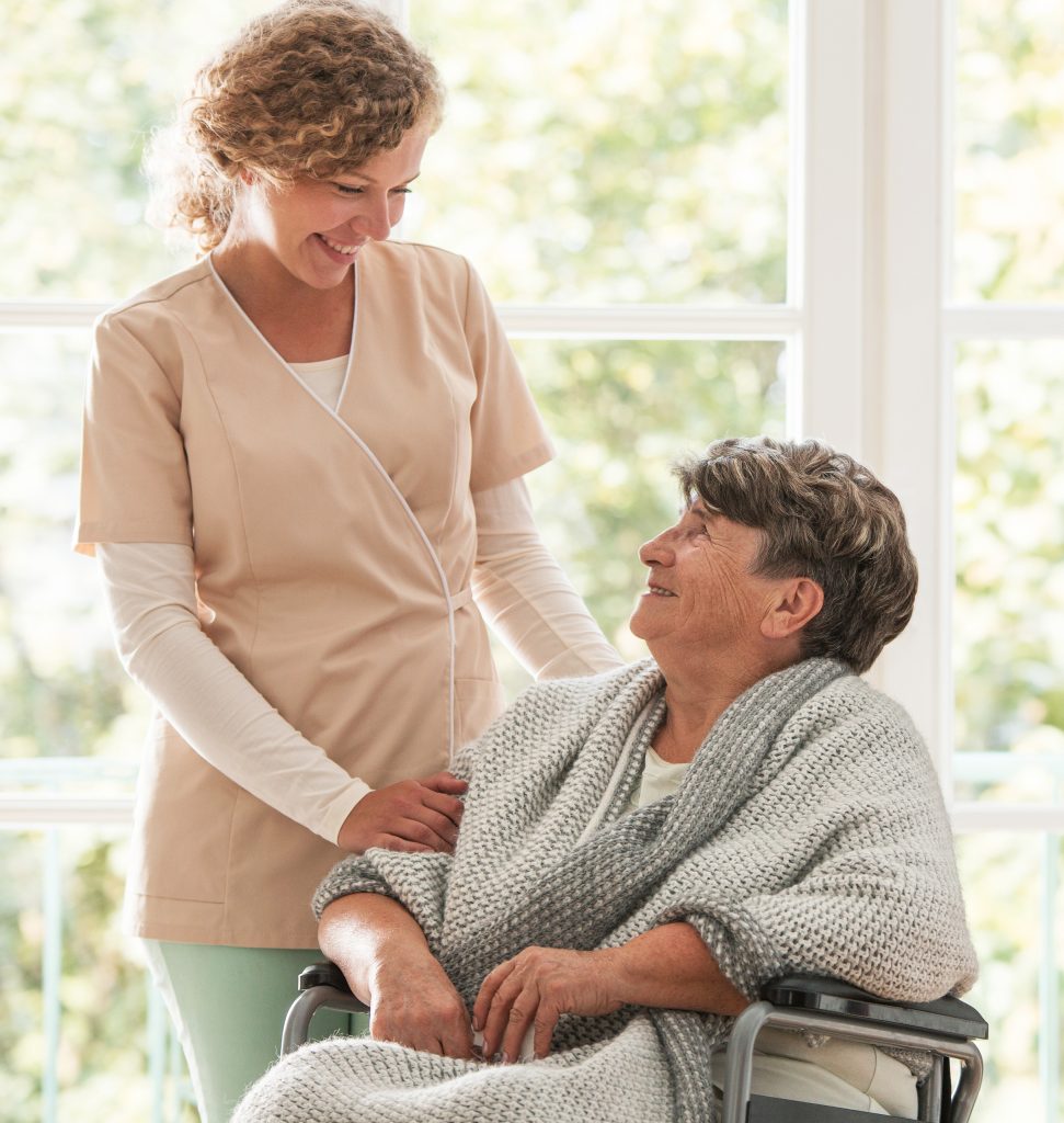a caretaker looks down and smiles at a resident in a wheelchair, while touching her arm in a comforting manner